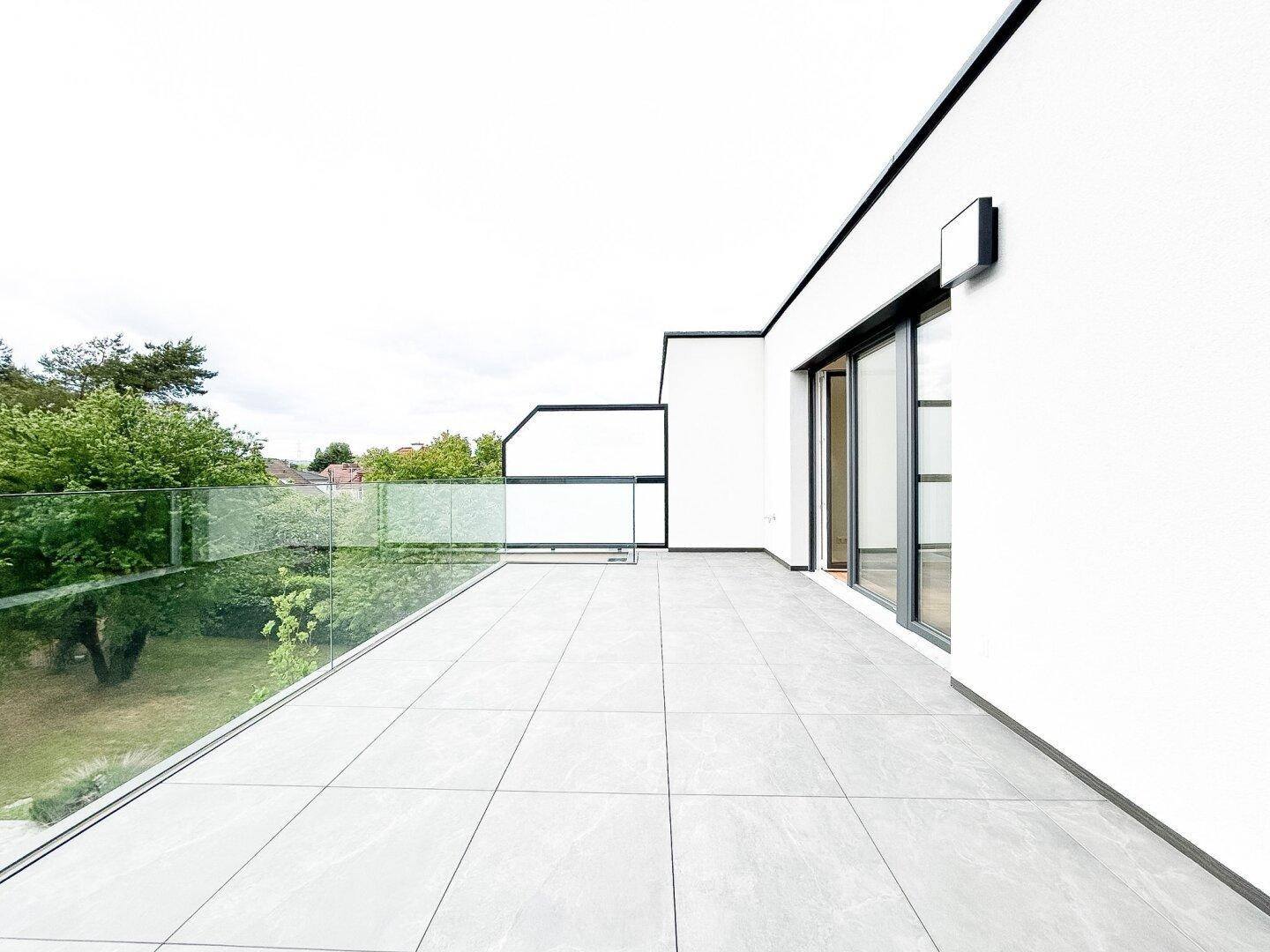 Geräumige, leere Terrasse mit modernen Fliesen und Glasgeländer, bereit zur individuellen Gestaltung.