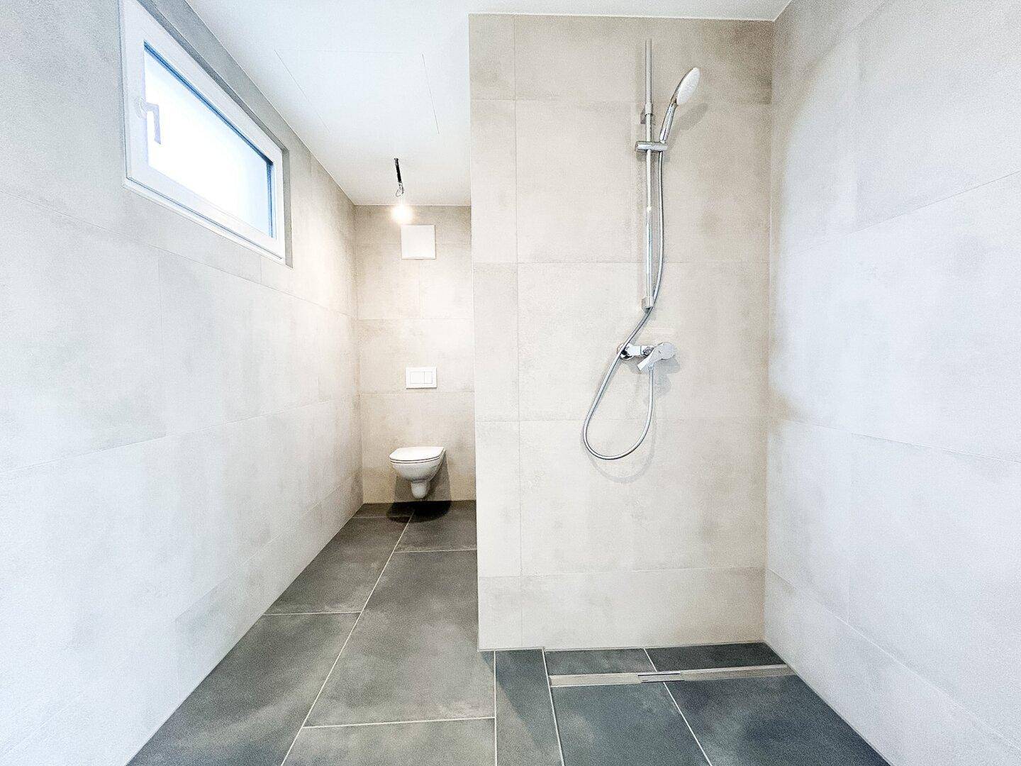 Modernes Badezimmer mit Toilette, ebenerdiger Dusche und hellgrauen Wand- und Bodenfliesen.