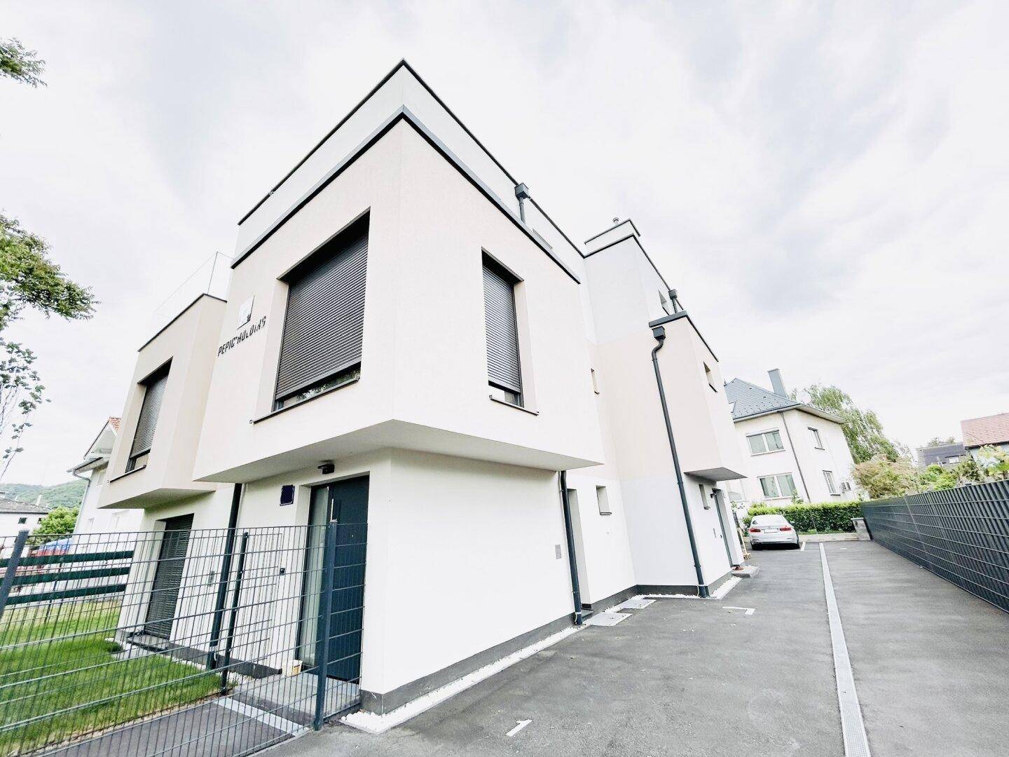 Moderne weiße Fassade eines Neubaus mit klaren Linien, schwarzen Akzenten und Rollläden.