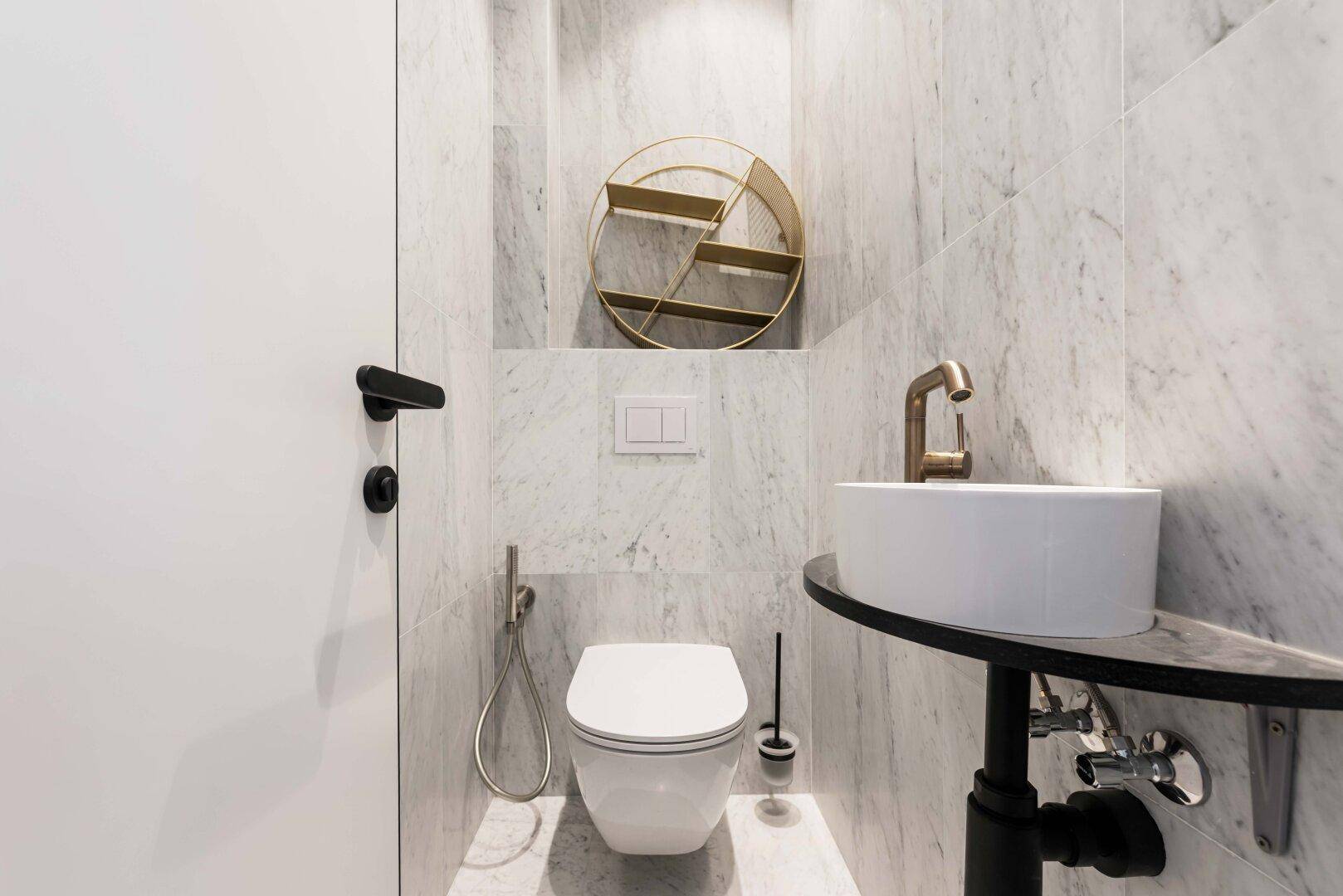 Modernes Gäste-WC mit Marmorfliesen, wandhängendem WC und stilvollem Waschbecken mit goldener Armatur.