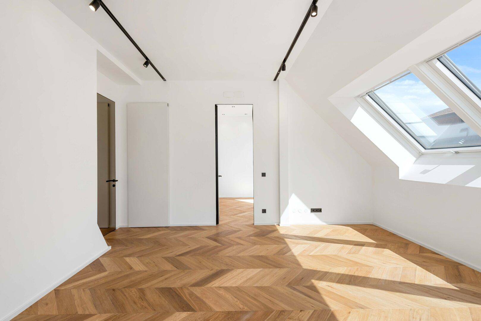 Heller Dachgeschossraum mit Fischgrätparkett, Dachfenstern und modernen, rahmenlosen Türen.