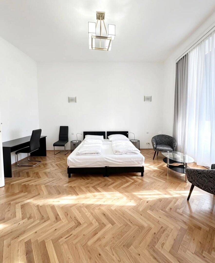 Helles Schlafzimmer mit Fischgrätparkett, Doppelbett und einem Schreibtisch am Fenster.