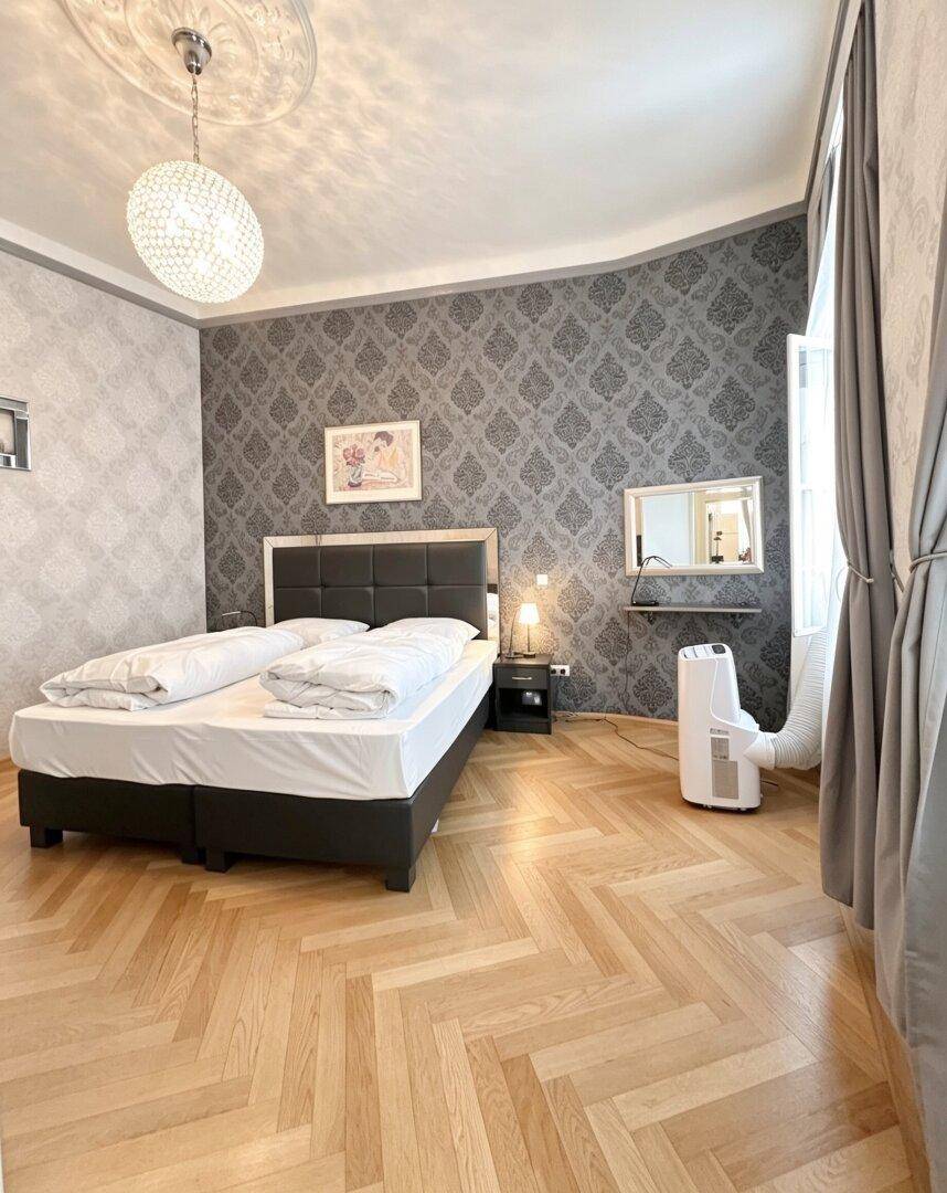 Schlafzimmer mit Fischgrätparkett, gemusterter Tapete und mobiler Klimaanlage.