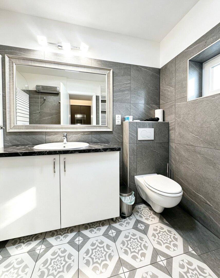 Modernes Badezimmer mit grauen Fliesen, Wand-WC und Fenster für Tageslicht.