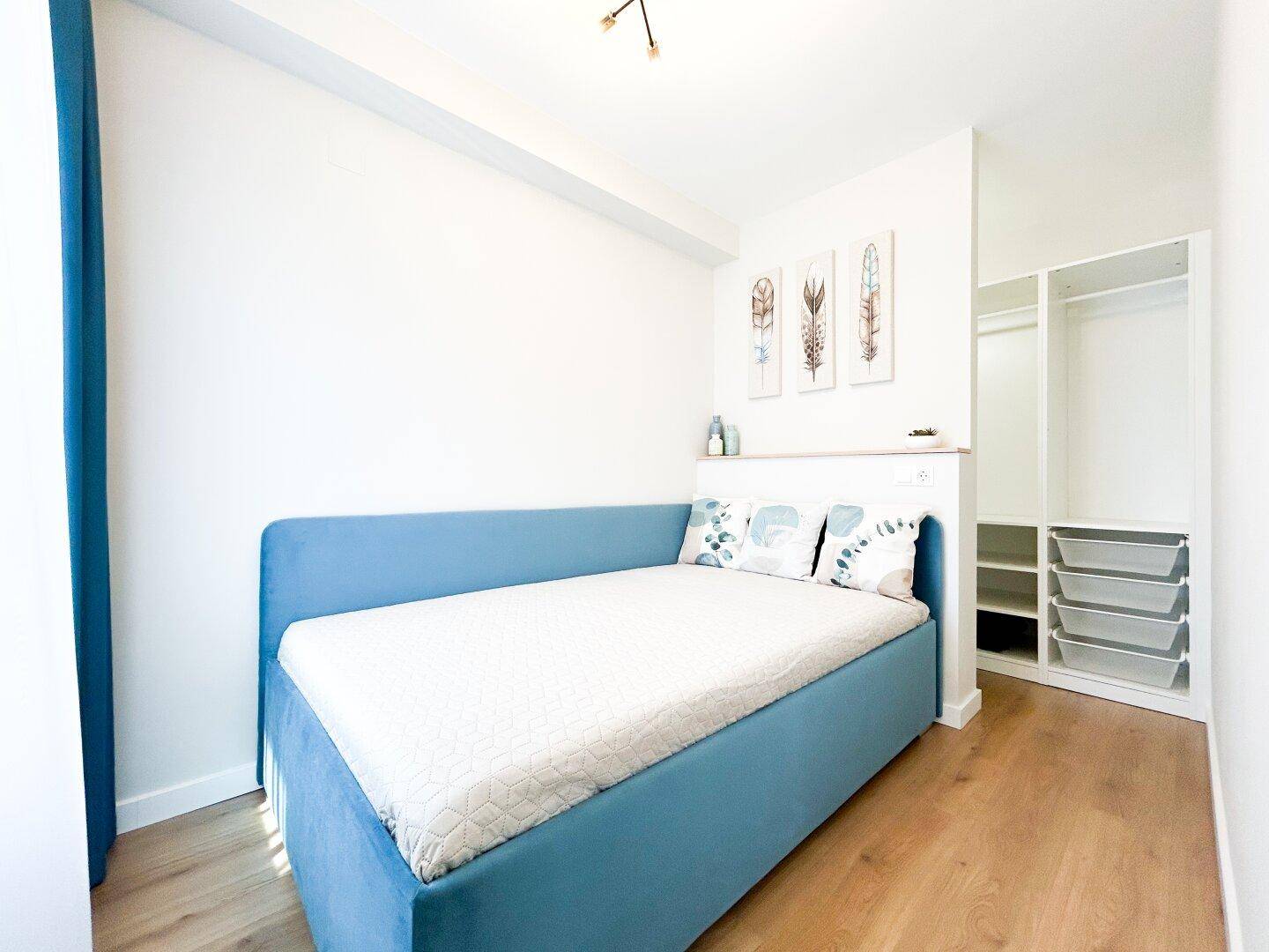 Helles Schlafzimmer mit blauem Bett, Holzboden und offenem Kleiderschrankbereich.