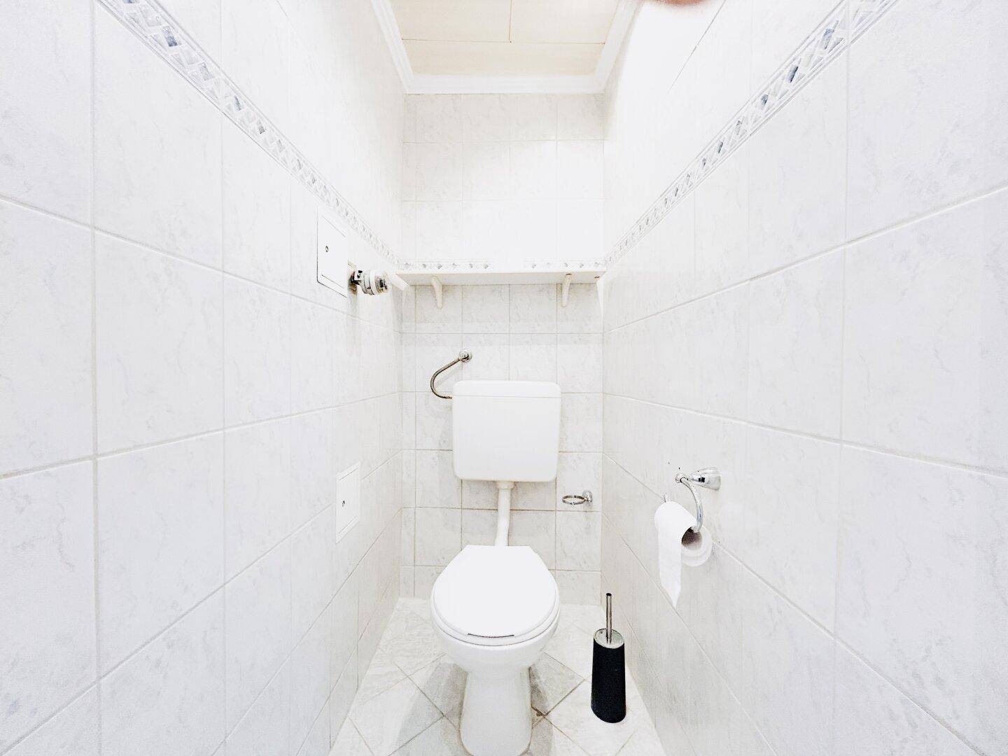 Separates WC mit weißen Fliesen an Wänden und Boden, ausgestattet mit einer Toilette.