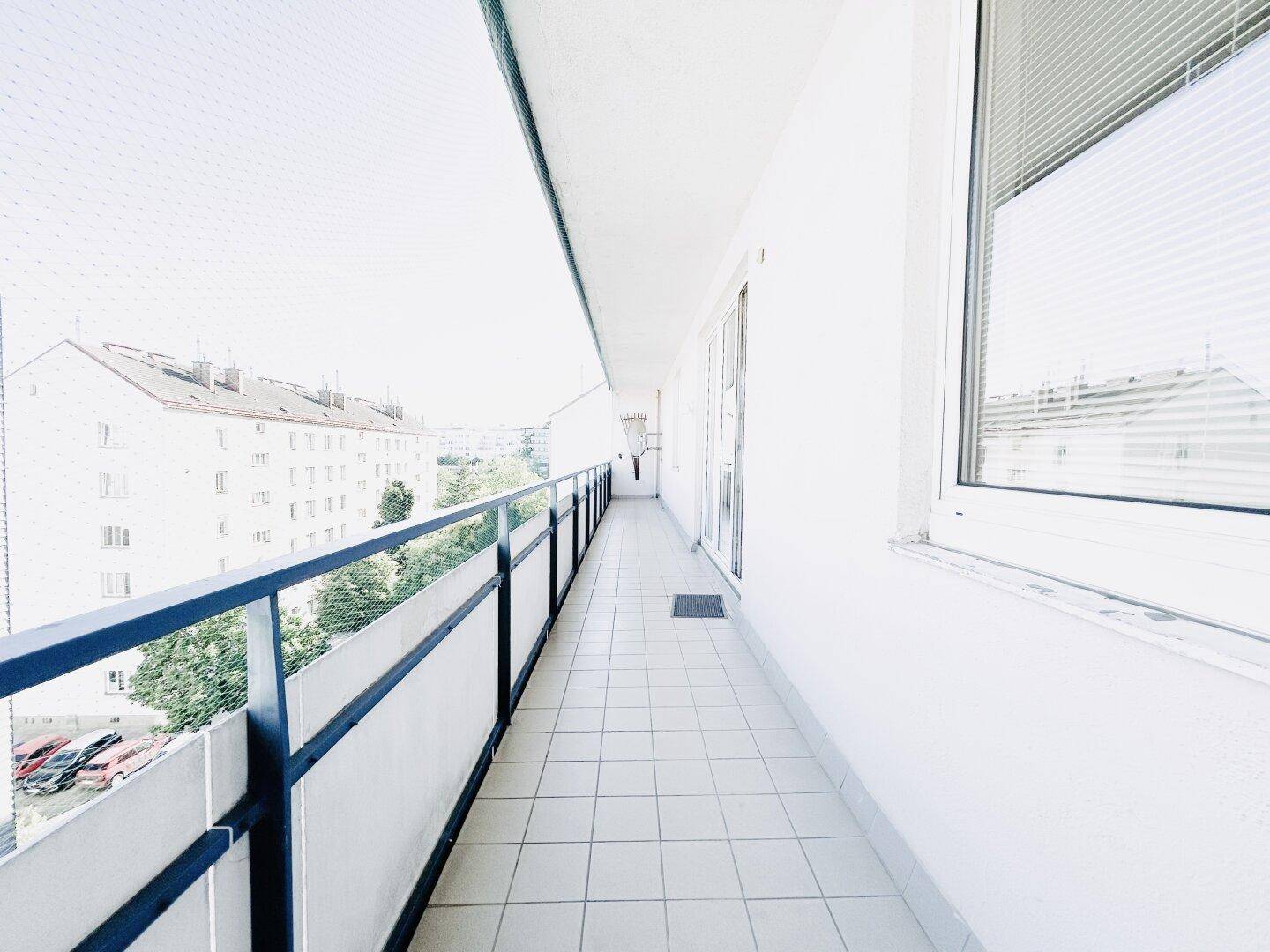 Langer Balkon mit Fliesenboden und Blick auf die umliegenden Gebäude und Bäume.