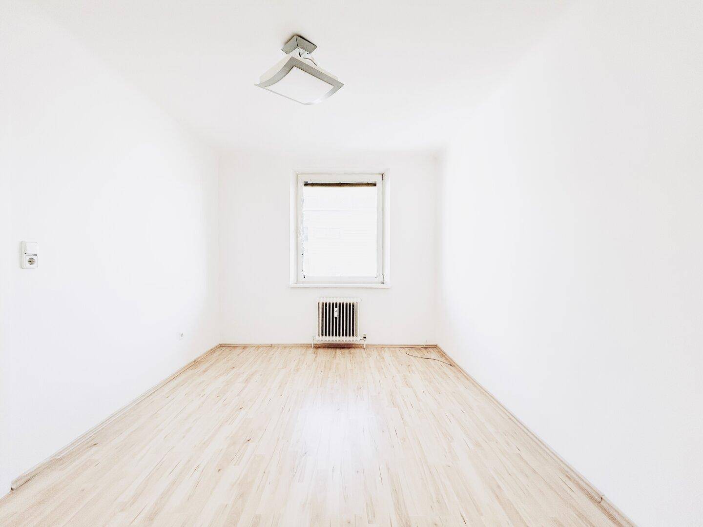Leeres Schlafzimmer mit hellem Laminatboden und einem Fenster, das viel Tageslicht hereinlässt.