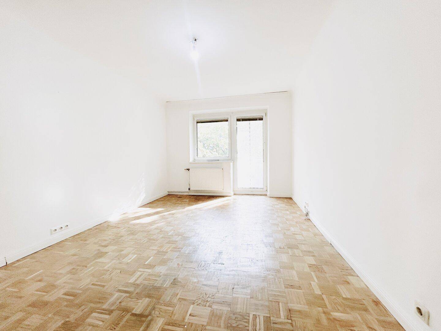 Sonniger, leerer Raum mit Parkettboden und Zugang zu einem möglichen Balkon oder einer Terrasse.