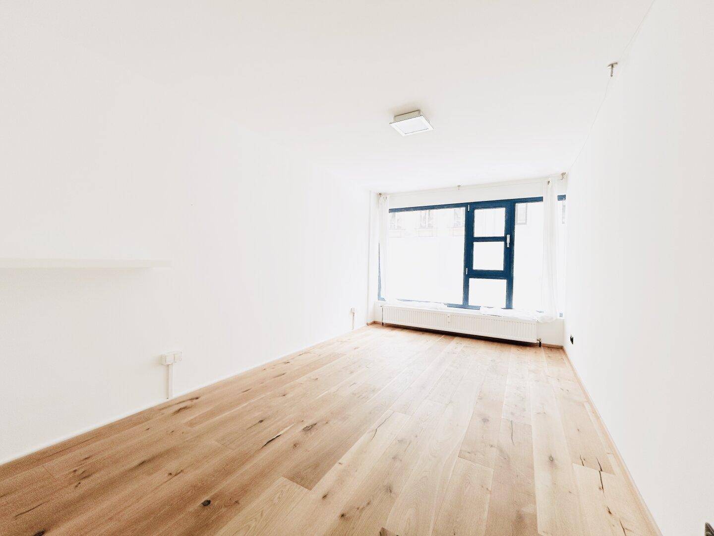 Leerer, heller Raum mit Holzboden und großem Fenster, bereit zur individuellen Gestaltung.