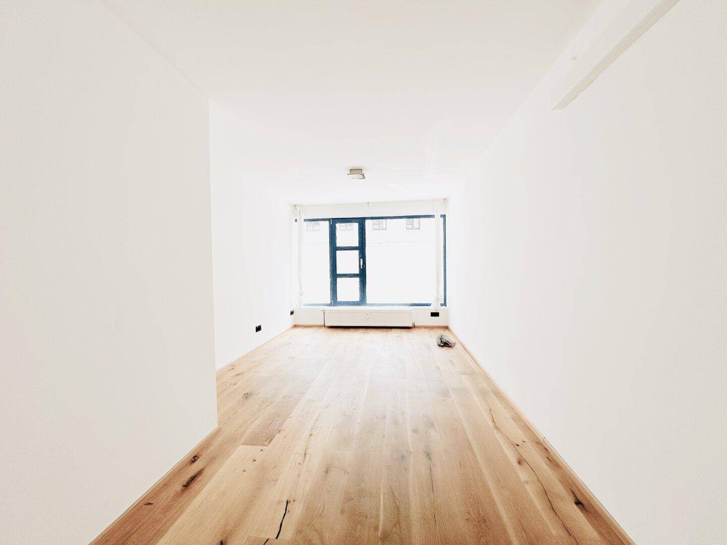 Leerer, heller Raum mit Holzboden und großem Fenster, das viel Tageslicht hereinlässt.