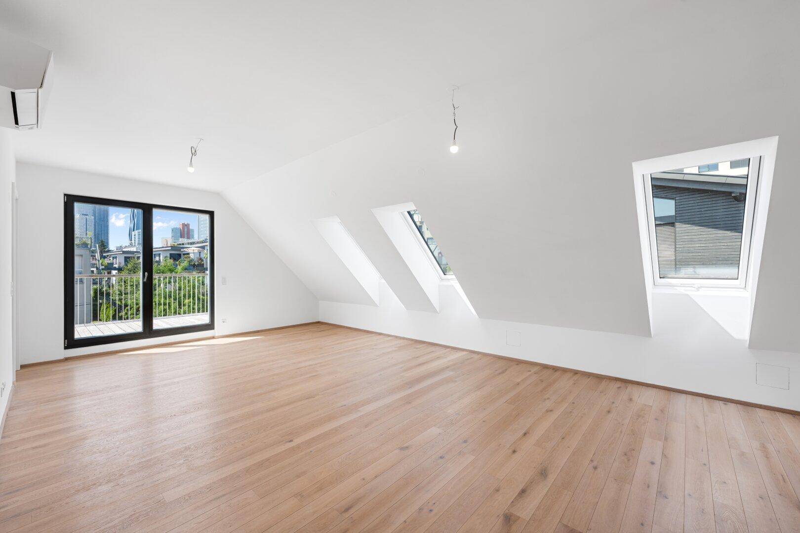 Heller, leerer Raum mit Holzboden, Dachschrägen und Zugang zu einem kleinen Balkon.
