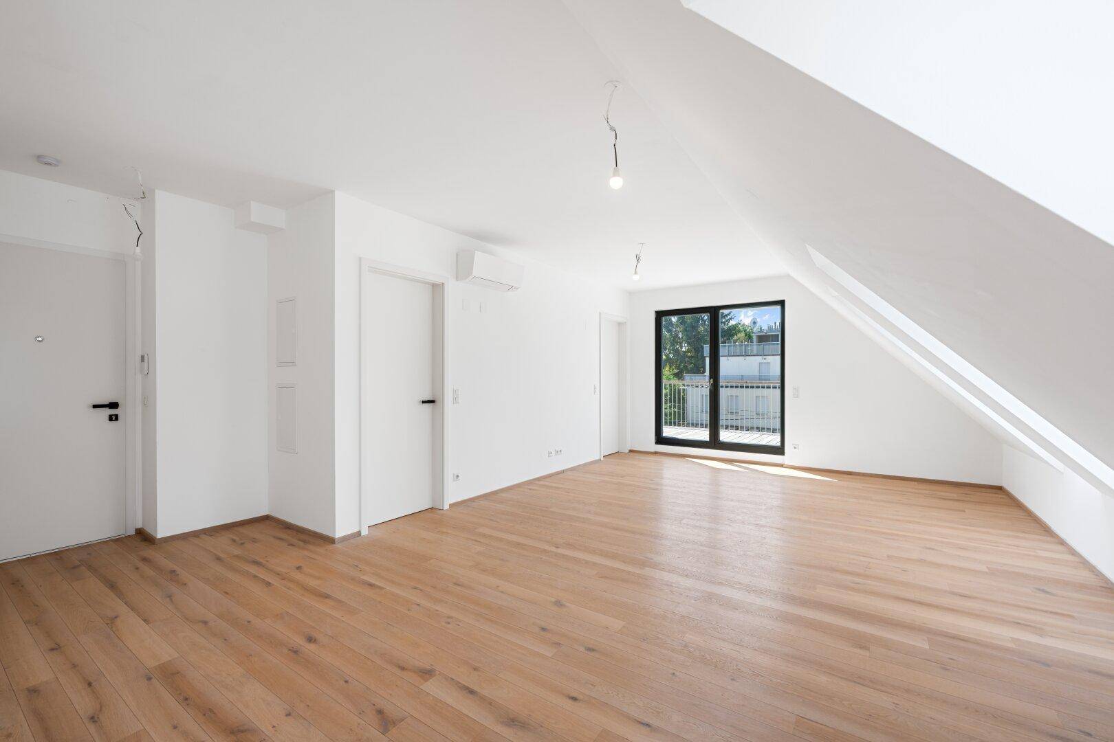 Heller Wohnbereich mit Holzboden, Dachschrägen und Zugang zu einem kleinen Balkon.