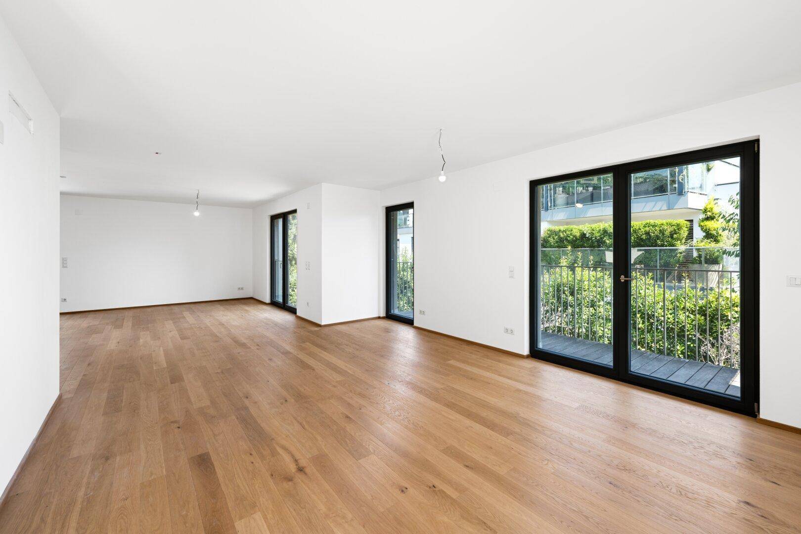 Helles, leeres Wohnzimmer mit Eichenparkettboden und großen Fenstern mit Zugang zum Balkon.