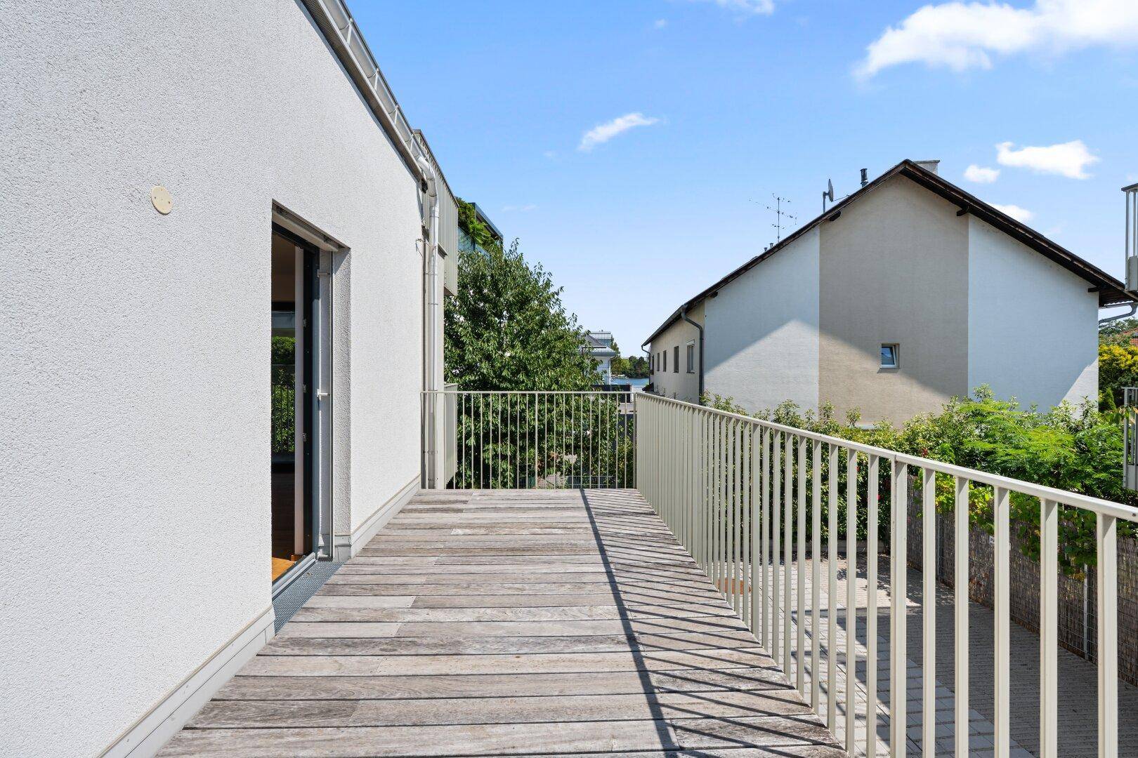 Langer Balkon mit Holzboden und Metallgeländer, umgeben von grünen Bäumen.