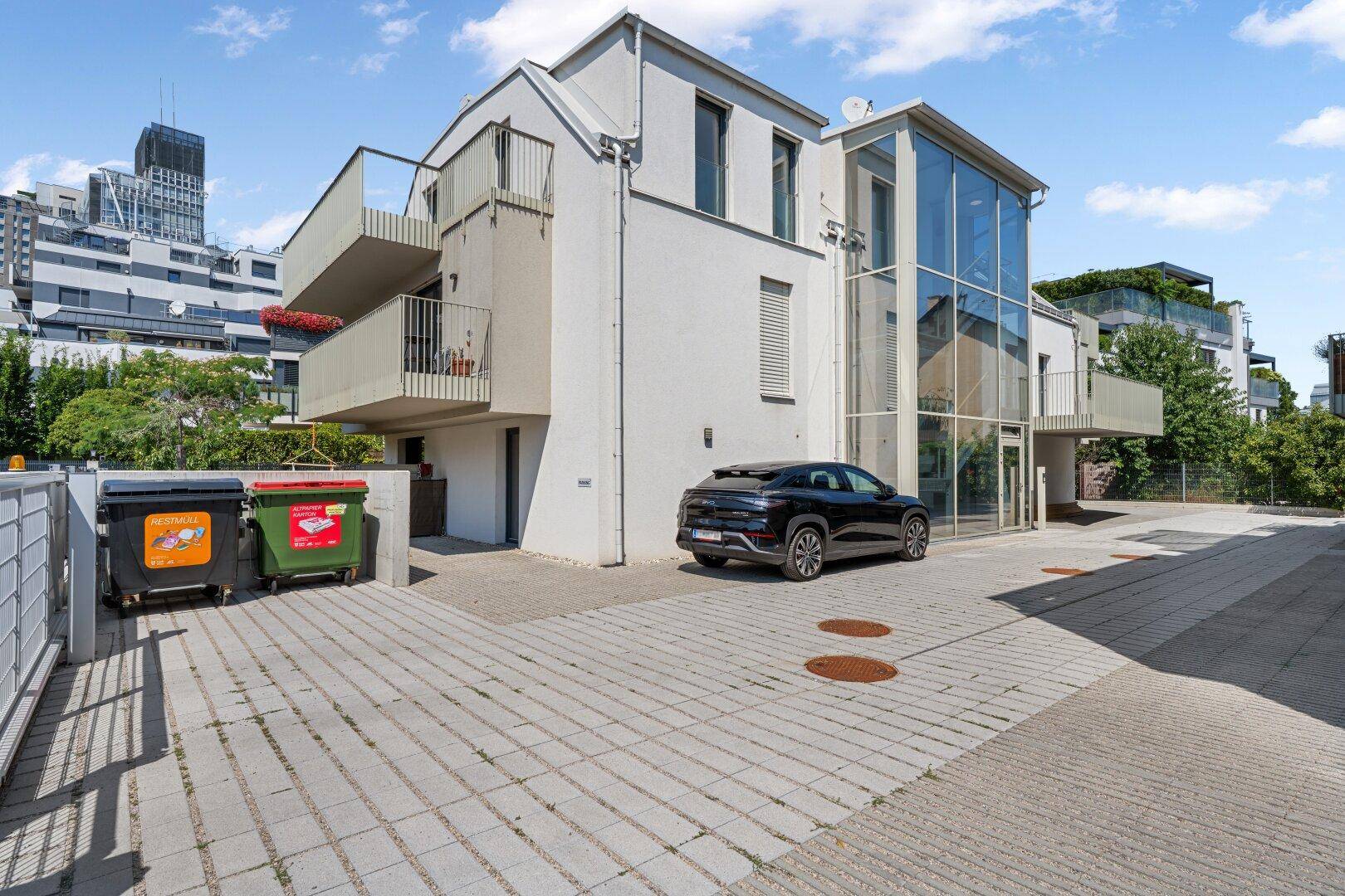 Modernes Gebäude mit Balkonen, Glasfassade und gepflastertem Parkplatz.