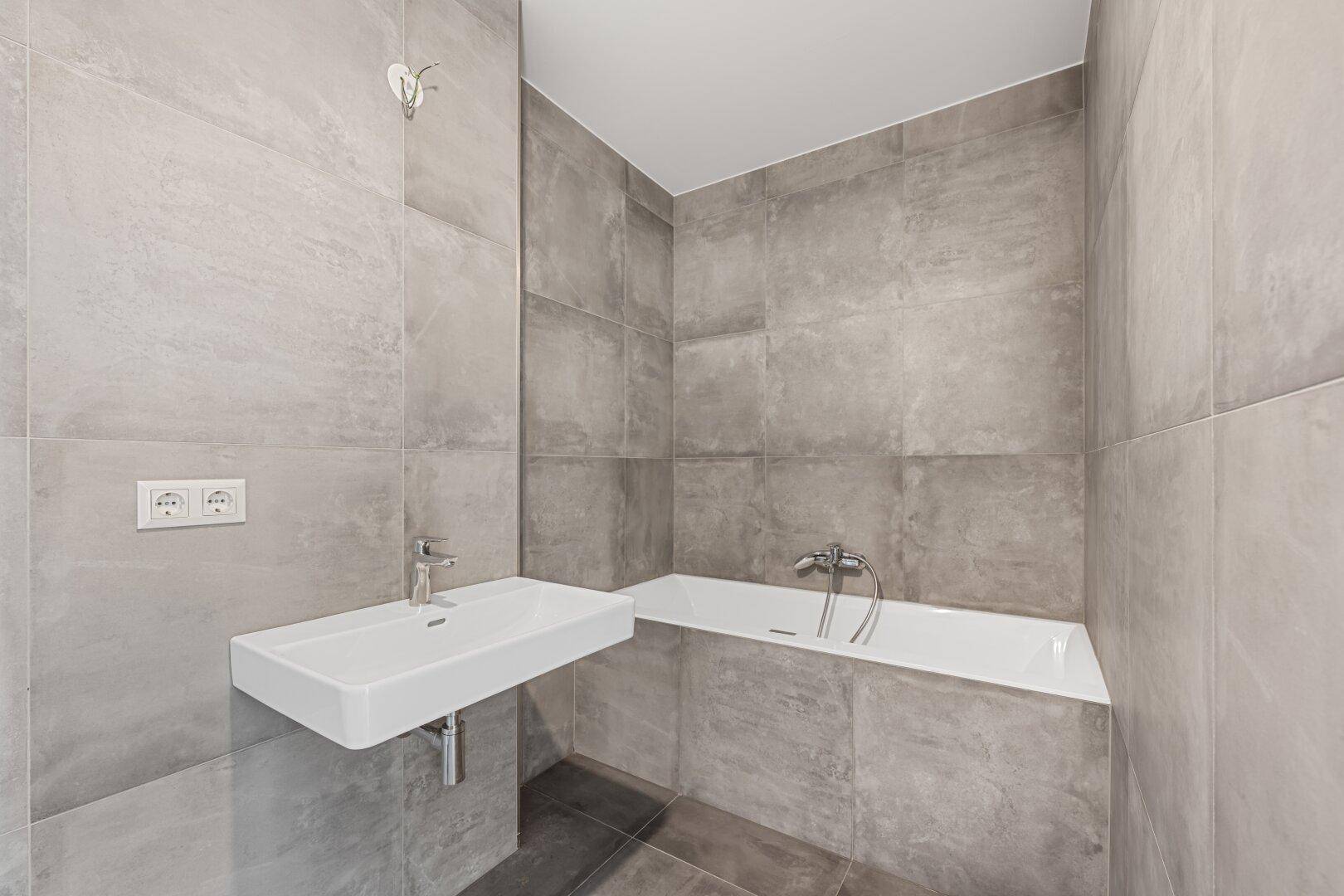 Modernes Badezimmer mit grauen Fliesen, integrierter Badewanne und schwebendem Waschbecken.