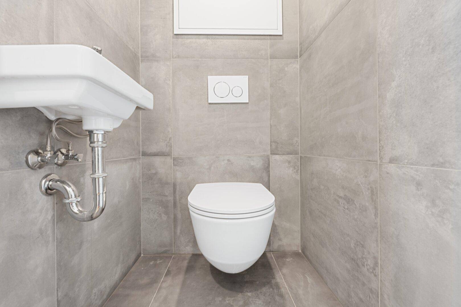 Modernes Gäste-WC mit grauen Fliesen, wandhängender Toilette und kleinem Waschbecken.