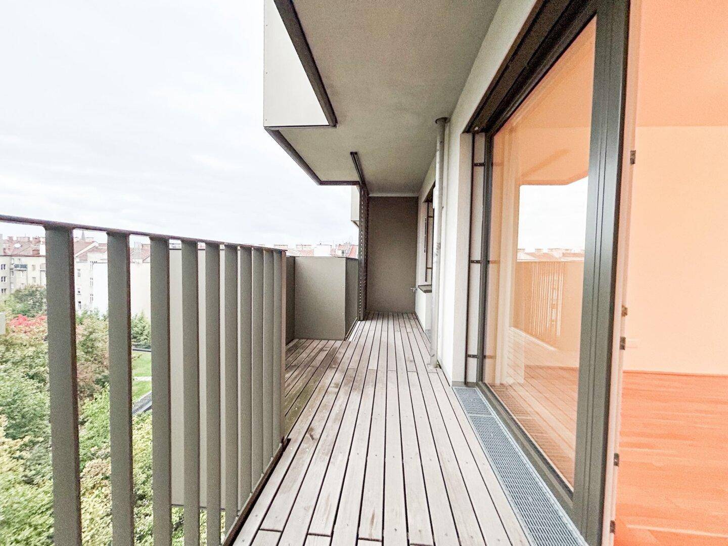 Langer, leerer Balkon mit Holzboden und Blick auf die umliegenden Gebäude und Bäume.