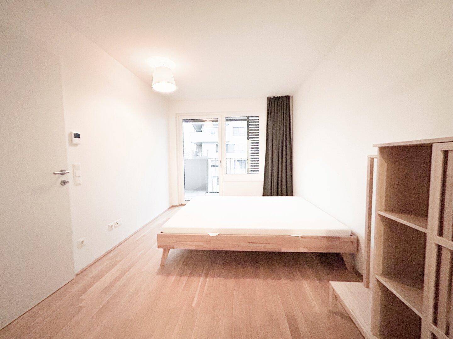 Helles Schlafzimmer mit Holzbettrahmen, Parkettboden und Zugang zum Balkon.