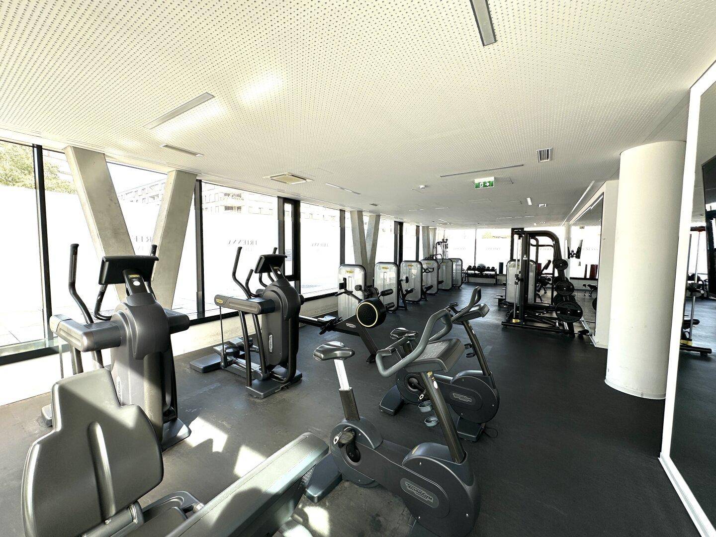 Gut ausgestatteter Fitnessraum mit verschiedenen Trainingsgeräten und großen Fenstern.