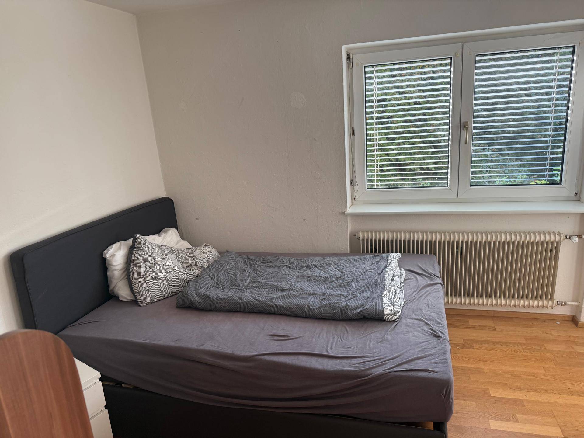 Gemütliches Schlafzimmer mit Bett, Fenster mit Jalousien und Heizkörper unter dem Fenster.