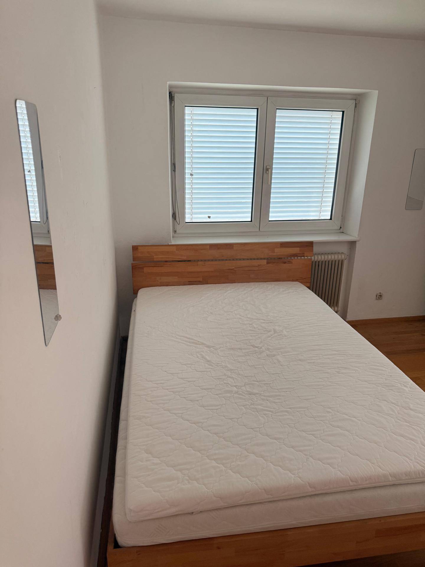 Helles Schlafzimmer mit Holzbett, Fenster mit Jalousien und Heizkörper unter dem Fenster.