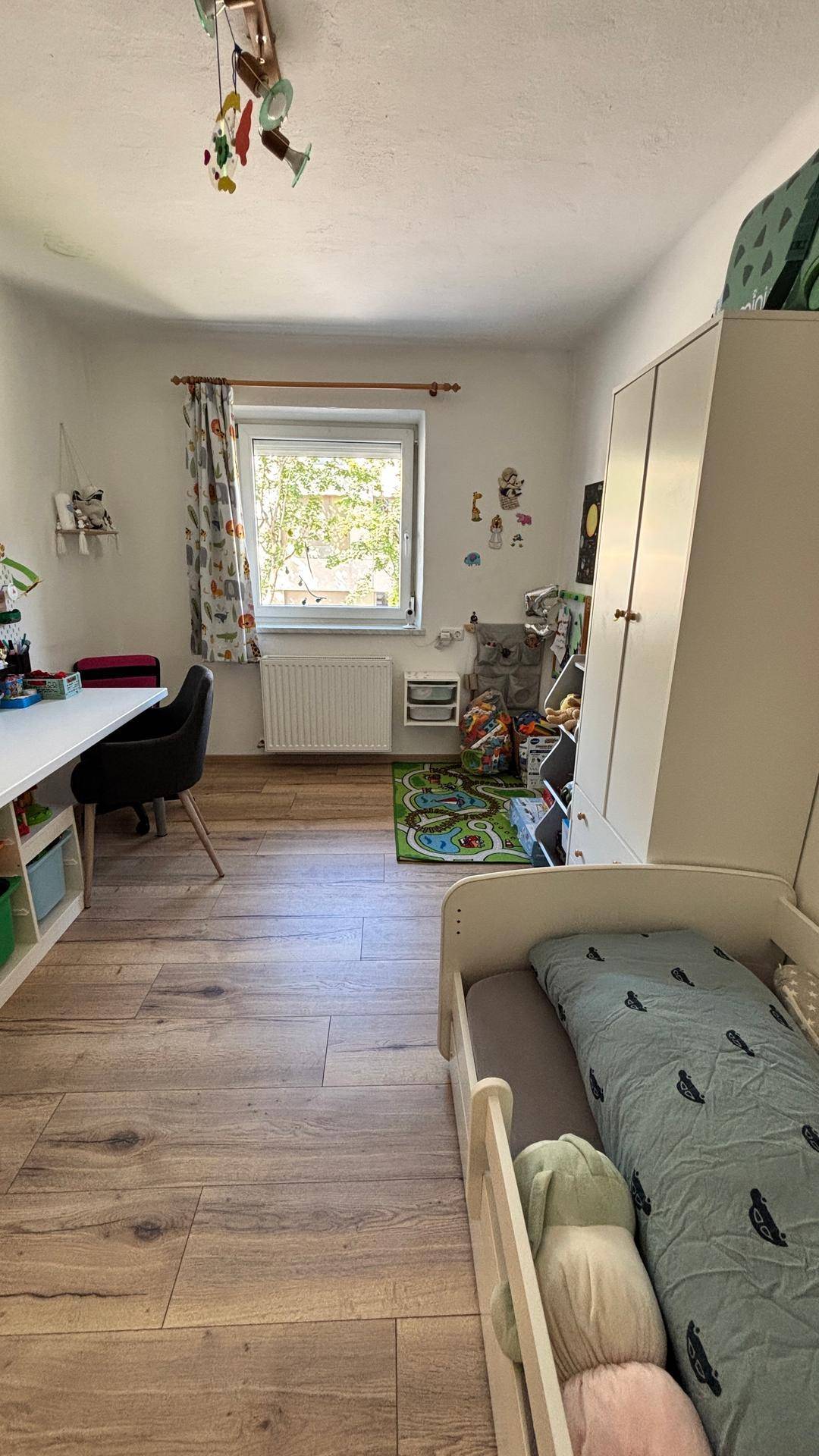 Helles Kinderzimmer mit Bett, Schreibtisch, Kleiderschrank und Blick ins Grüne durch das Fenster.