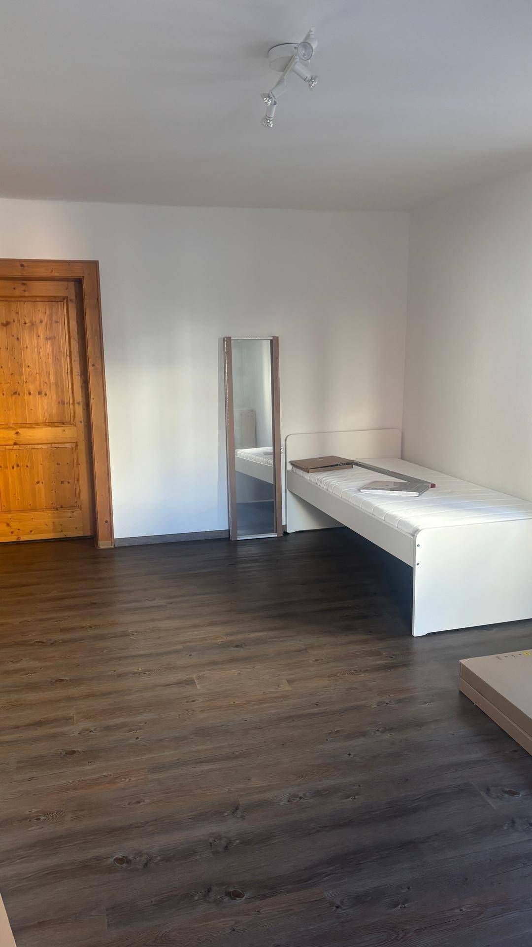 Schlichtes Schlafzimmer mit einem Einzelbett, einem großen Spiegel und dunklem Laminatboden, bereit zur Einrichtung.