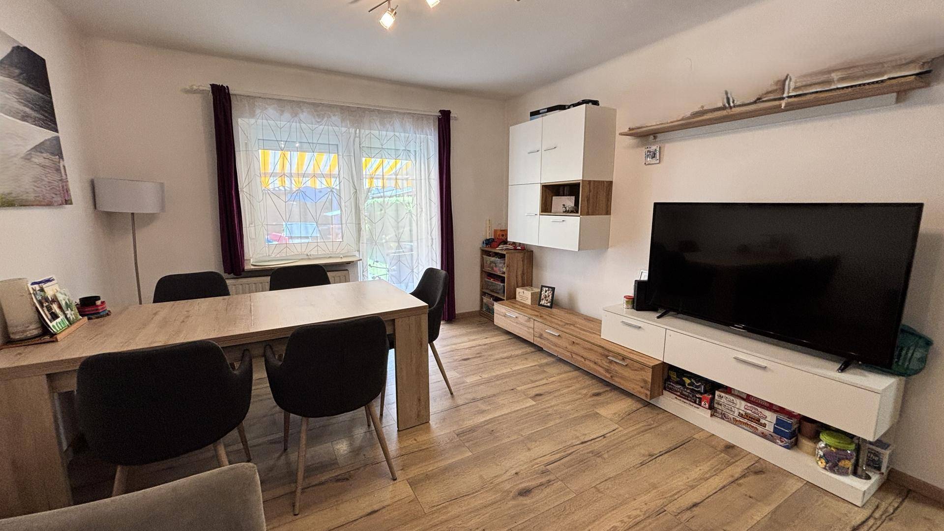 Gemütlicher Wohnbereich mit TV-Möbeln, Esstisch und Blick auf den Garten durch das Fenster.