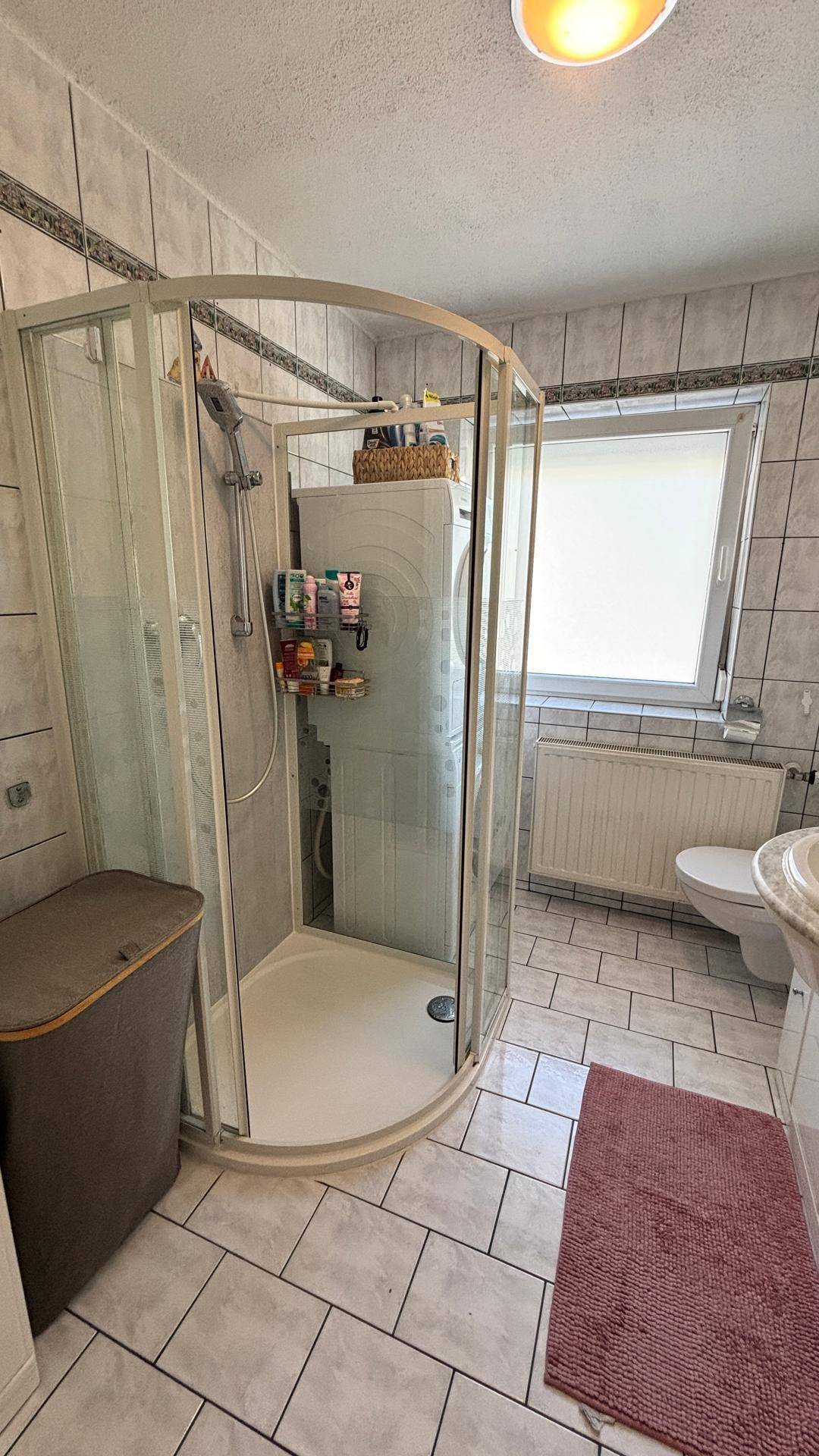 Badezimmer mit Eckduschkabine, WC und Waschbecken, hell gefliest und mit Fenster.