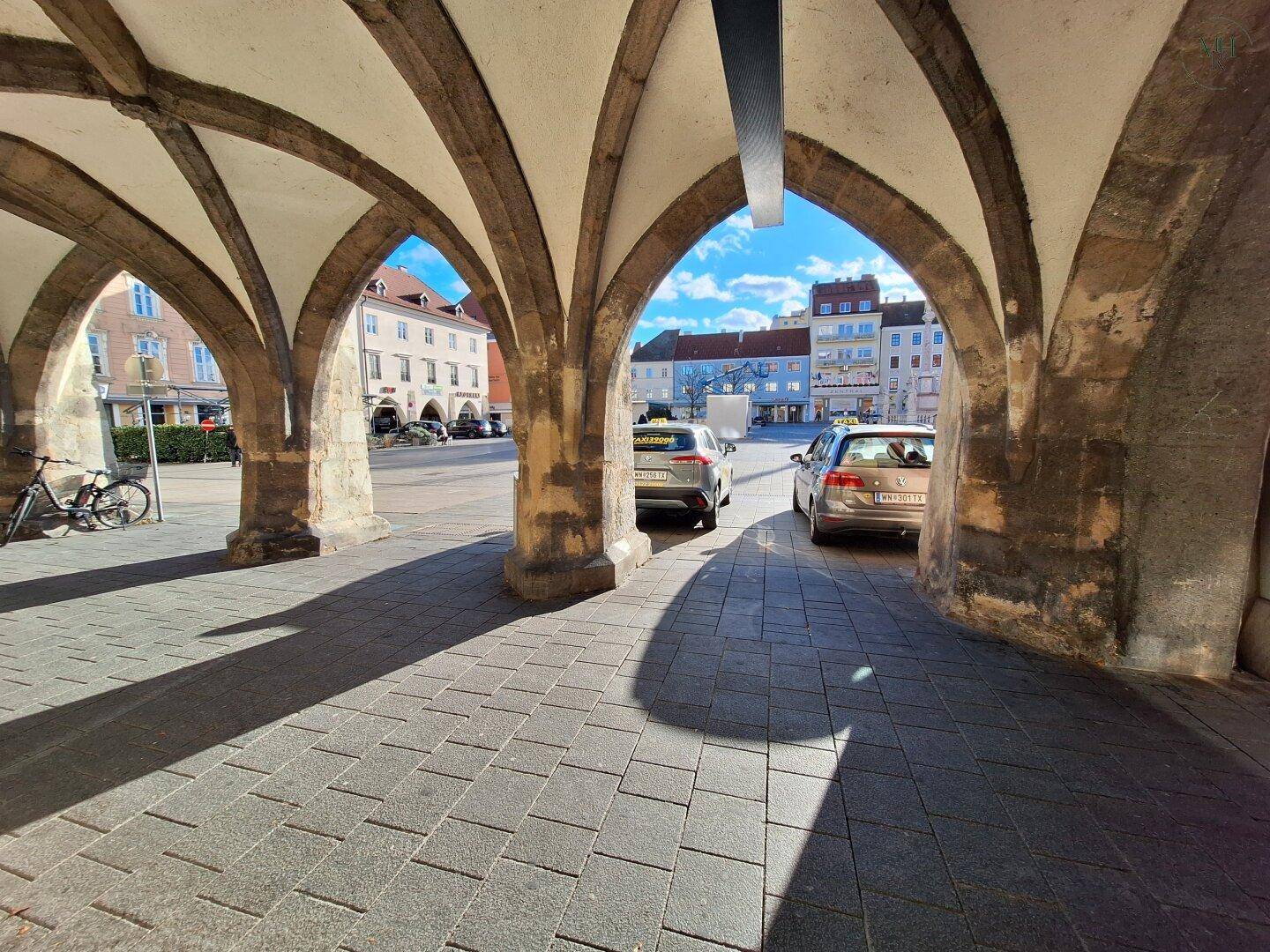 Blick von einer historischen Arkade auf einen sonnigen Stadtplatz mit geparkten Autos und traditionellen Gebäuden im Hintergrund.