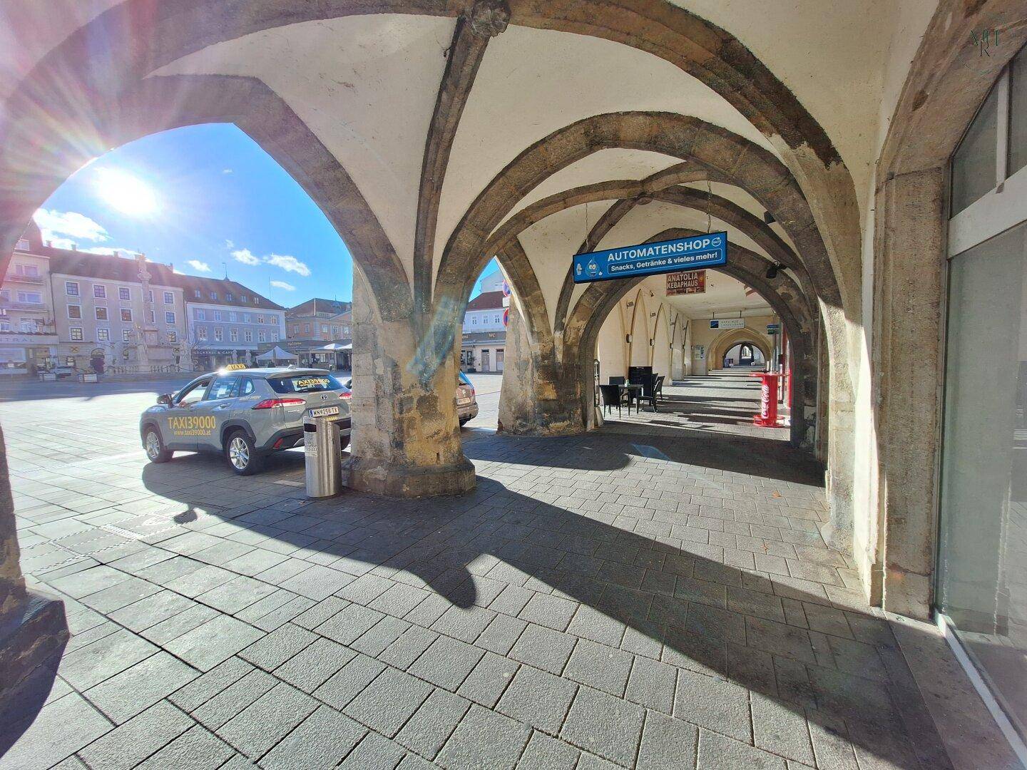 Sonniger Blick unter einer historischen Arkade auf einen belebten Stadtplatz mit einem Hinweisschild für einen Automatenladen.