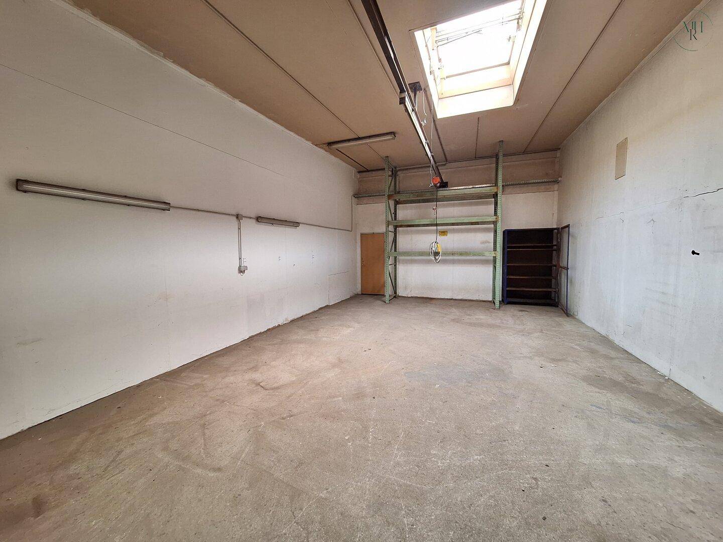 Leerer Lagerbereich mit Betonboden, Oberlicht und langen Leuchtstoffröhren an der hohen Decke.