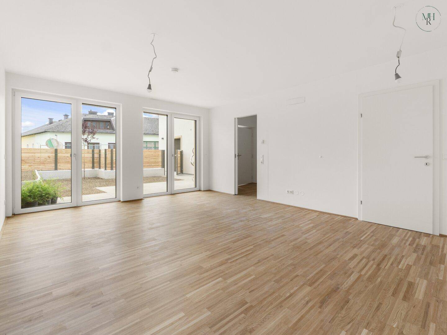 Heller, leerer Wohnraum mit Holzboden und großen Fenstern, die Zugang zum Gartenbereich bieten.