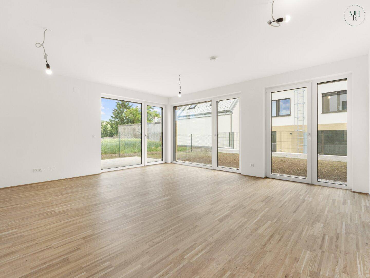 Heller, leerer Wohnraum mit großen Fensterfronten, Holzboden und Zugang zum Garten.