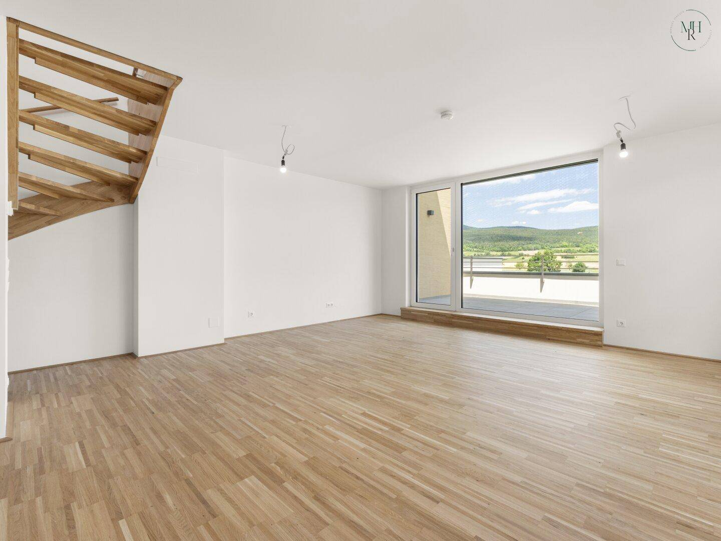 Helles, leeres Wohnzimmer mit Holzboden, großen Fenstern und Zugang zur Terrasse mit Landschaftsblick.