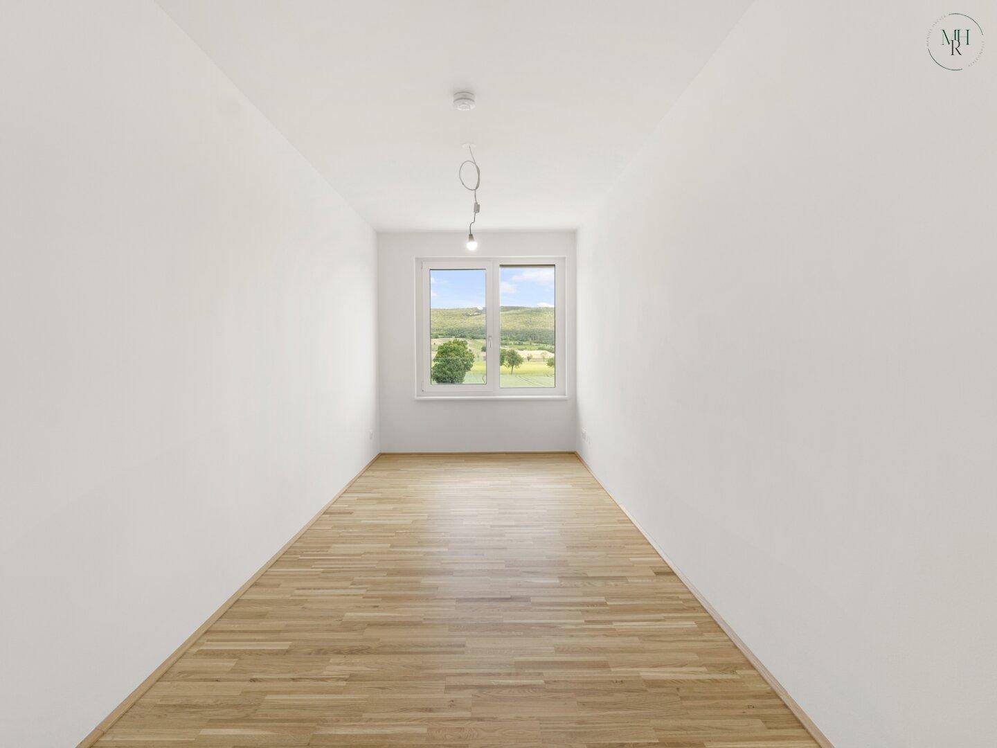 Leeres Zimmer mit Holzboden und einem Fenster, das einen weiten Blick auf die grüne Landschaft bietet.