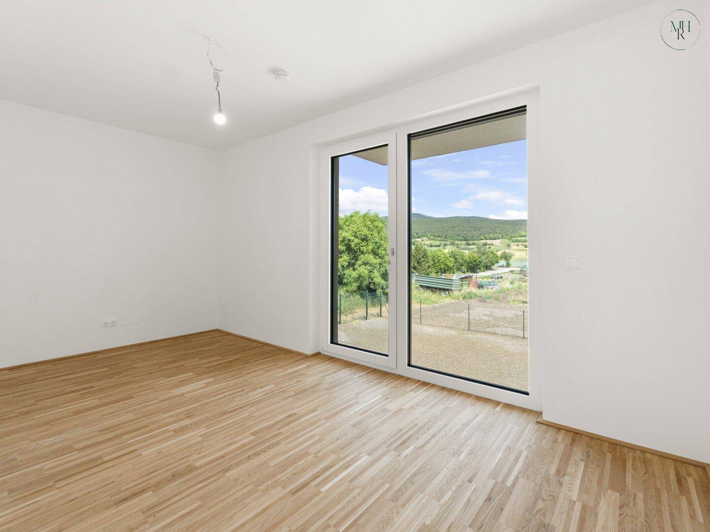 Helles Zimmer mit Holzboden und großer Schiebetür, die Zugang zu einem Außenbereich mit Landschaftsblick bietet.