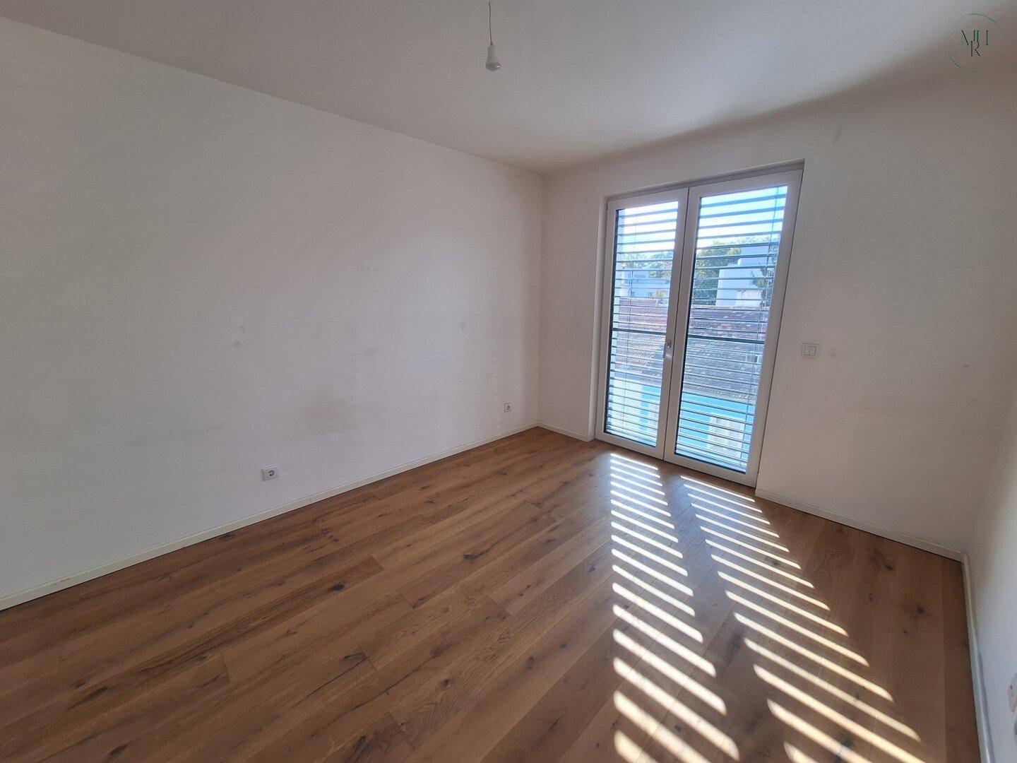 Helles Zimmer mit Holzboden und Fenster mit Jalousien, das viel Sonnenlicht hereinlässt.