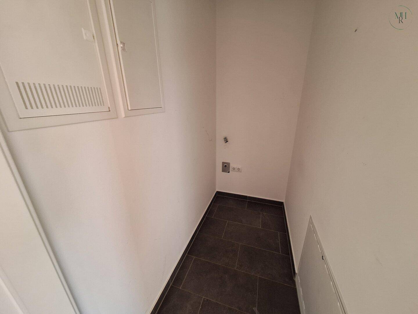 Praktischer Hauswirtschaftsraum mit dunklem Fliesenboden und Anschlüssen für eine Waschmaschine.