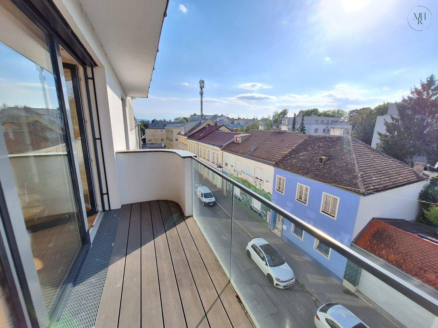 Moderner Balkon mit Holzdeck und Glasbrüstung, bietet einen weiten Blick über die Straße und Häuser.