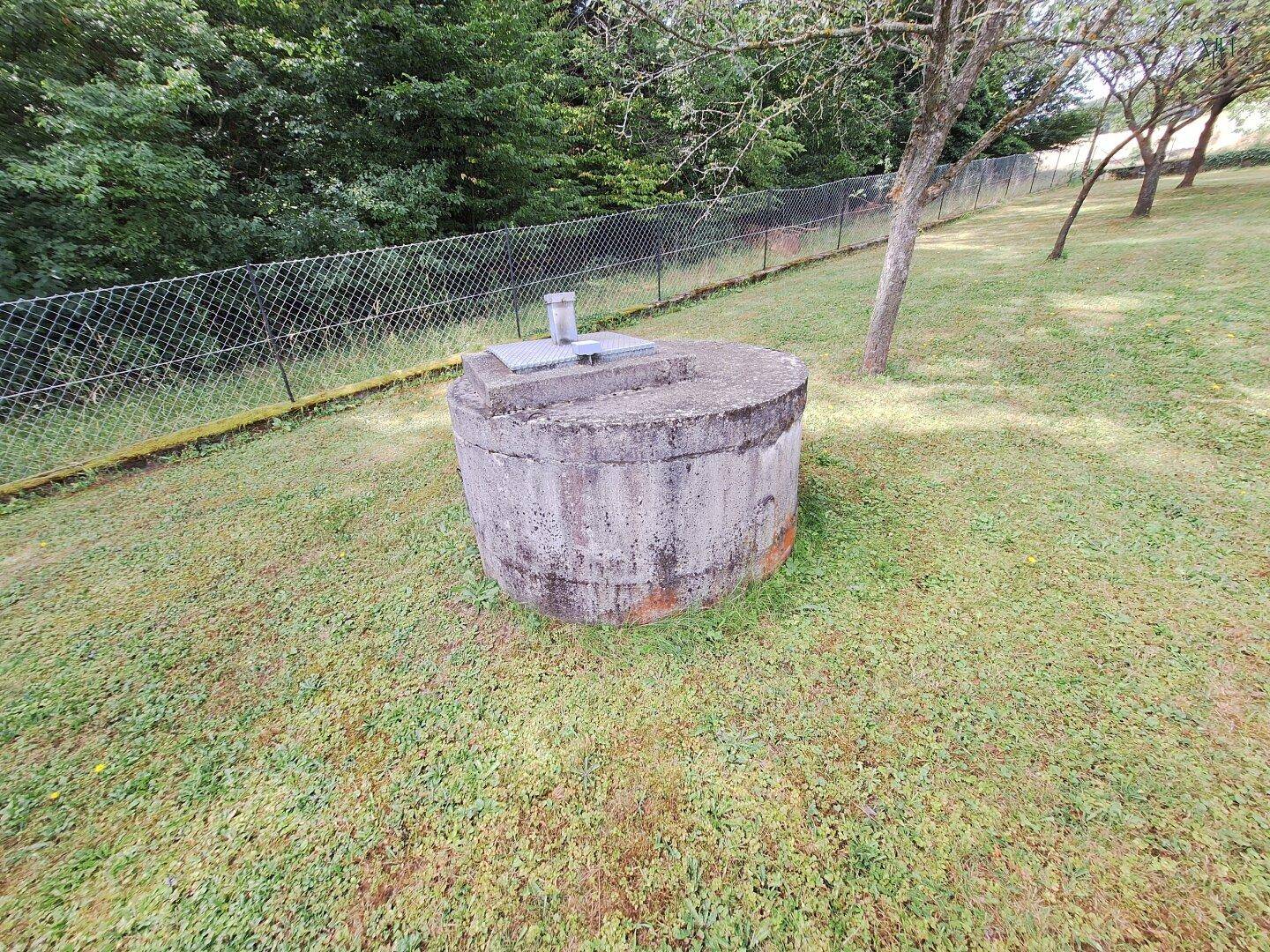 Betonbrunnen oder Zisterne im grünen Gartenbereich, umgeben von Rasen und einem Zaun.