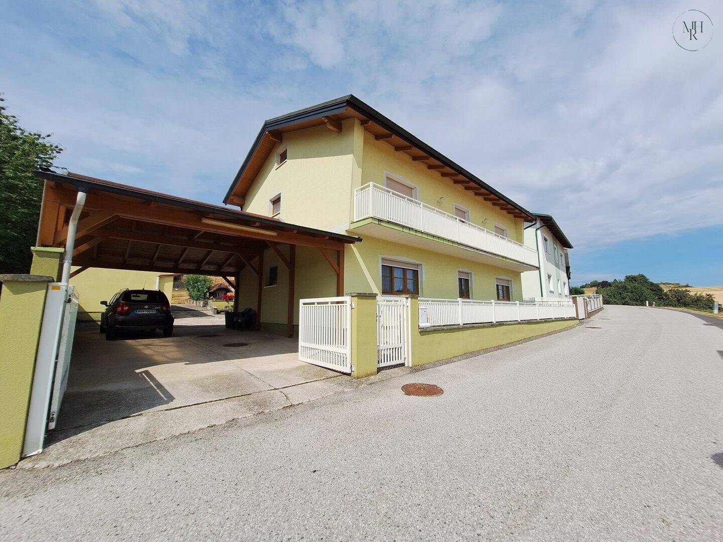 Ansicht eines gelben Hauses mit Balkonen und einem Carport von der Straße aus, umgeben von Grün.
