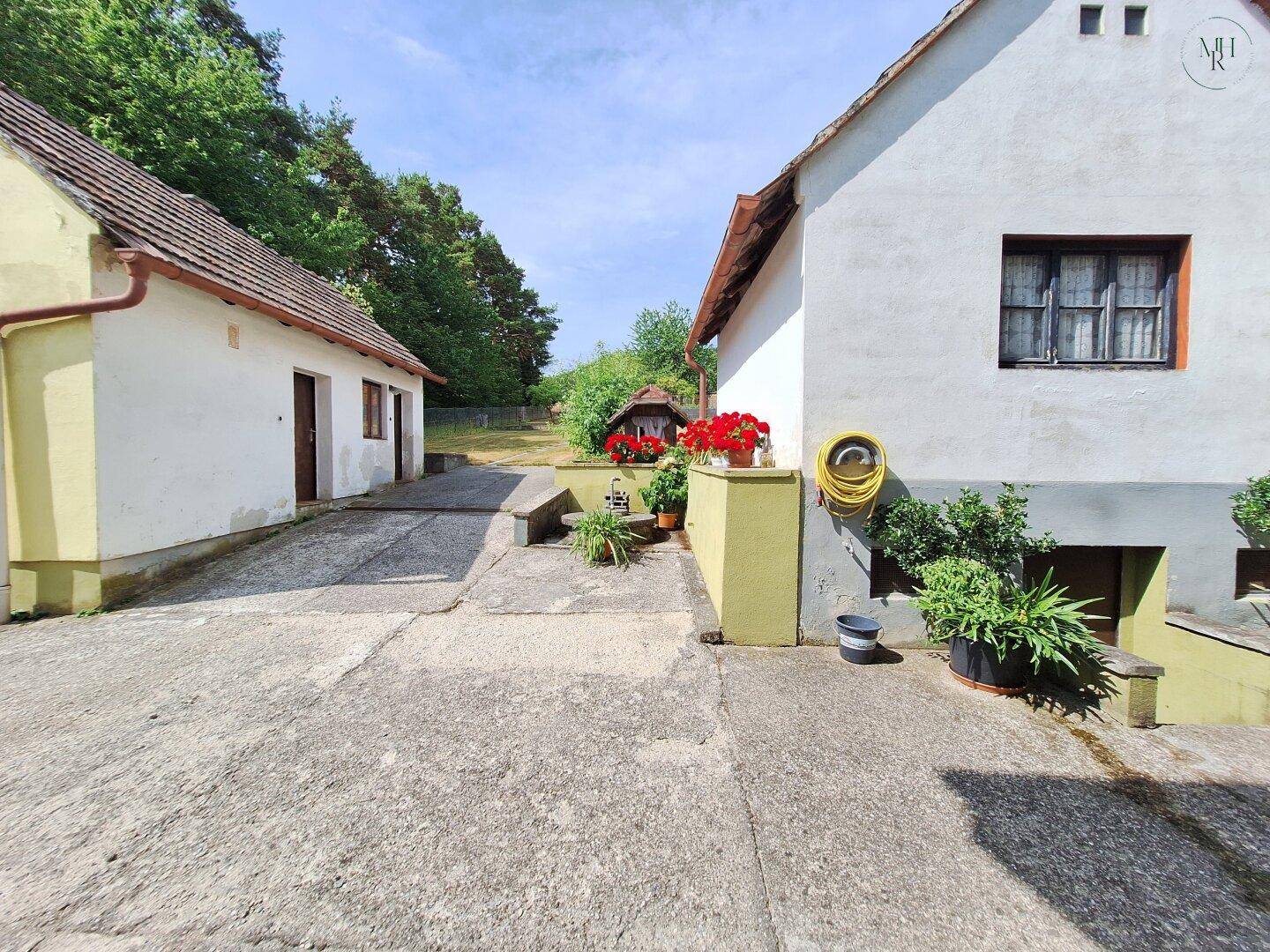 Gepflasterter Bereich zwischen zwei Gebäuden, der zu einem bewaldeten Hang und offener Natur führt.