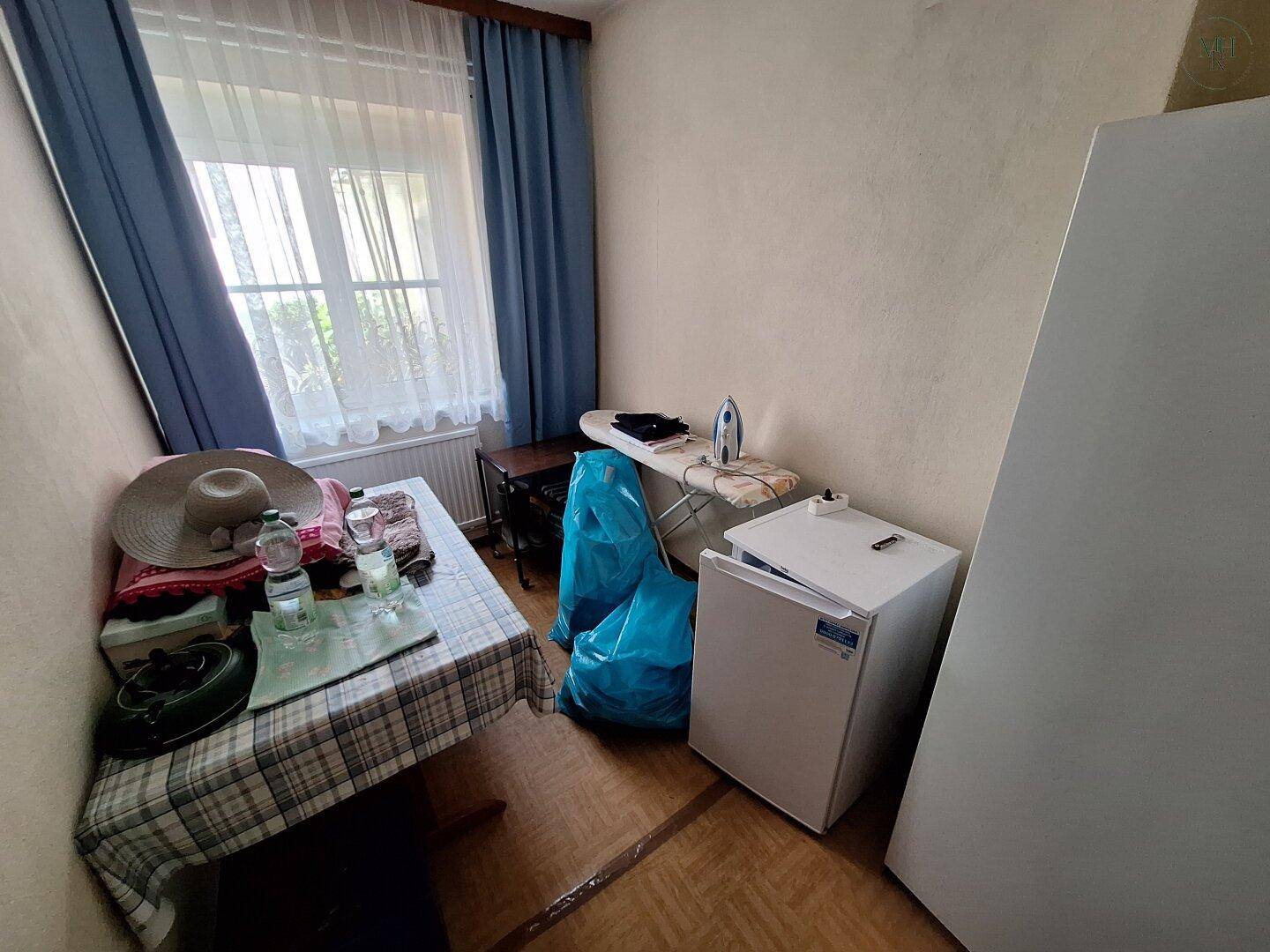 Kleiner Abstellraum mit Bügelbrett, Kühlschrank und einem Fenster mit blauen Vorhängen, praktisch eingerichtet.
