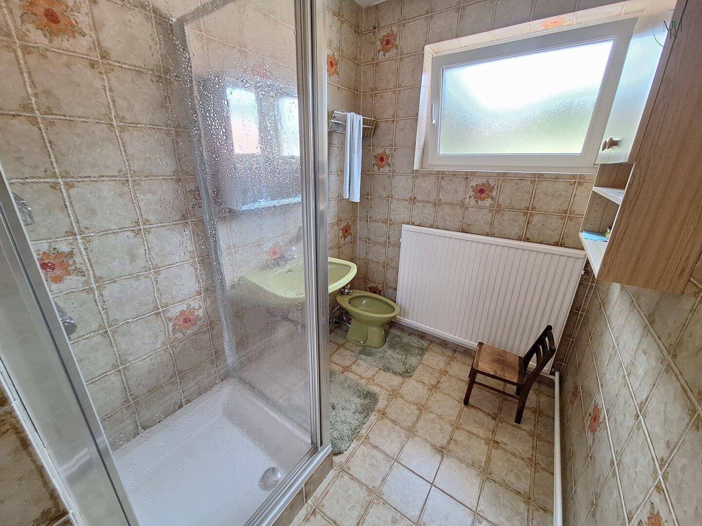 Badezimmer mit Duschkabine, WC und floralen Wandfliesen im älteren, aber gepflegten Stil.