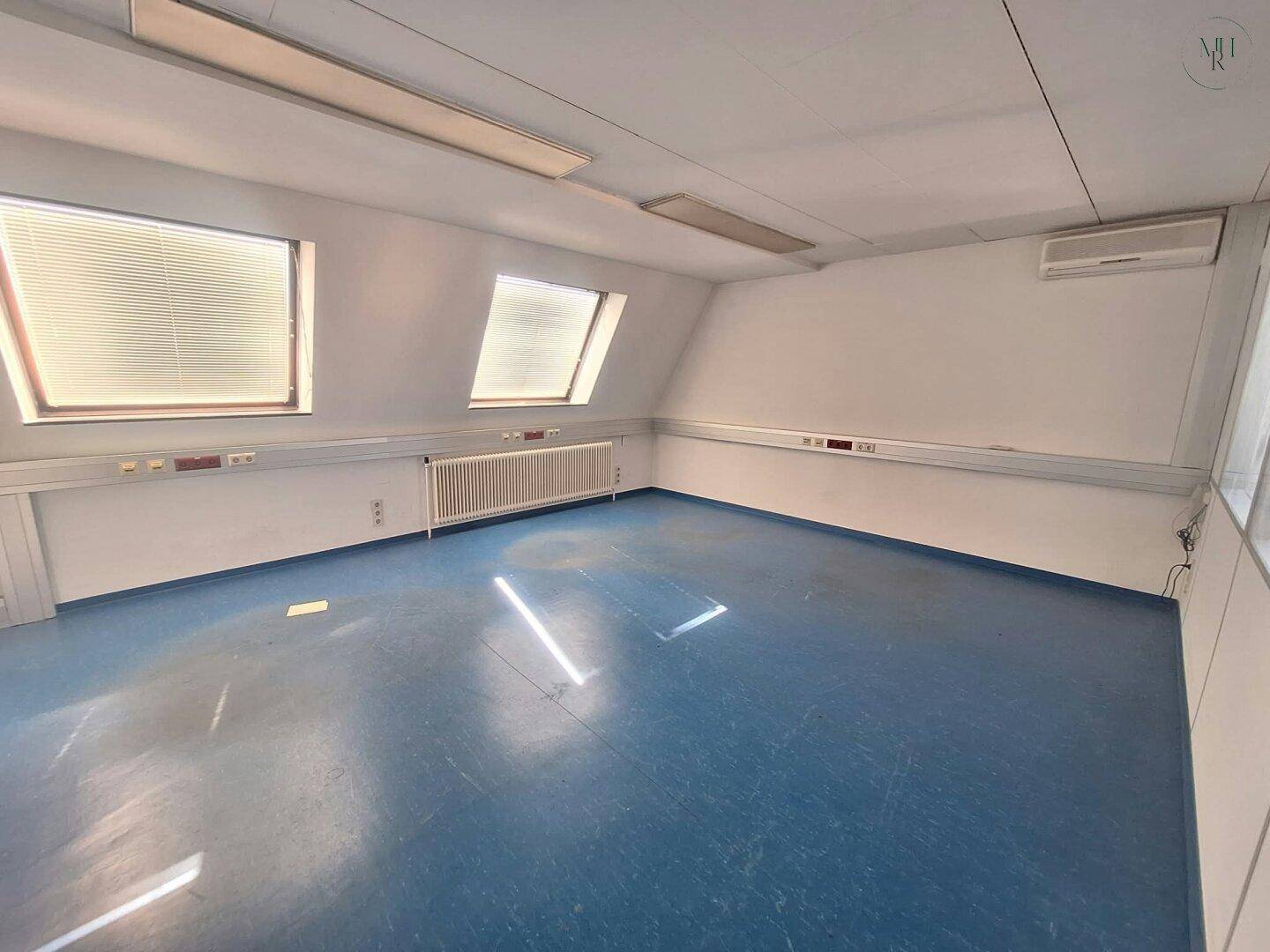 Geräumiger Büroraum mit blauer Bodenfläche, Dachfenstern und Klimaanlage an der Wand.