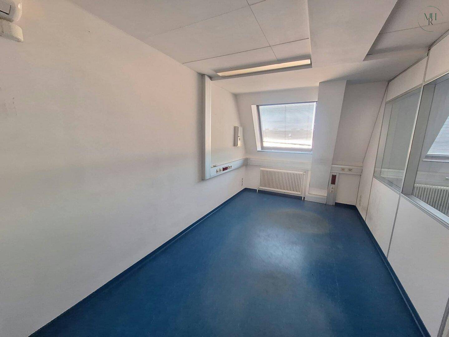 Büroraum mit schrägen Wänden, einem Dachfenster und Heizkörper, ausgestattet mit blauer Bodenfläche.