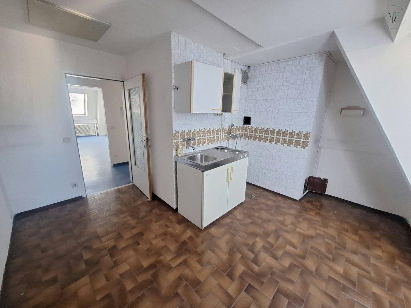 Raum mit kleiner Küchenzeile, gemustertem Boden und schräger Decke, angrenzend an einen weiteren Raum.